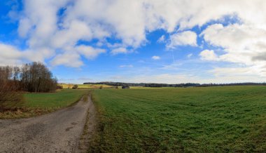 A dirt road leads out of the shade into the sun under blue sky near Geltendorf in Bavaria between meadows