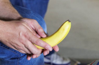 Beyaz adamın erkek eli bacaklarının arasında soyulmamış muz tutuyor.