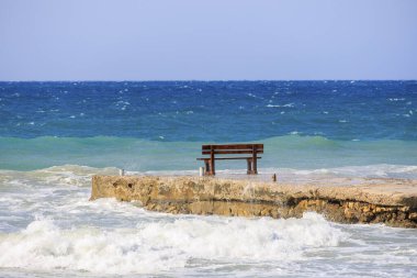 Dalgaların ortasında, Arillas kasabası yakınlarındaki Korfu adasında, mavi gökyüzü ve ağır denizlerin altında bir bank.