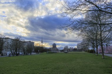 Gün batımında güneş ve bulutların dramatik bir karışımı televizyon kulesi ve Milbertshofen Almanya Münih 'teki Petuelpark' taki BMW şirket binası manzarası 16.3.2024
