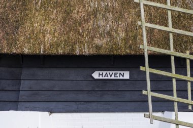 A signpost sign with the Dutch text Harbour on a dark painted wooden wall under a thatched roof of a polder windmill for draining the ditches in a residential area in Den Oever in the Netherlands, 24.3.2024 clipart