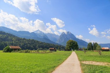 Garmisch-Partenkirchen yakınlarında güneşli bir günde mavi gökyüzü ve beyaz kümülüs bulutlarıyla Zugspitz kalabalığını gören bir patika yolu.