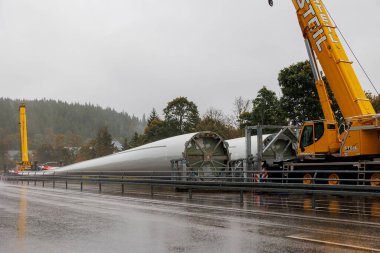 Kamyona monte edilmiş bir vinç Feldberg yolunun kenarında duruyor. 26 Eylül 2024 'te, Almanya, Feldberg' de, yağmur ve siste bir rüzgar türbininin iki pervanesinin yanında.