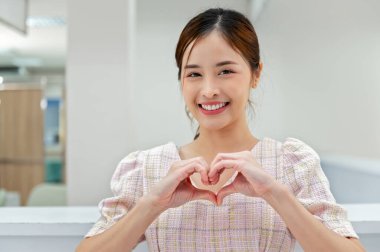 Güzel kadın gülümseyerek odada kalp şeklinde eller yapıyor.