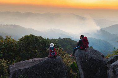 Bir adam ve bir kadın dağ sırasına bakan bir kayanın üzerinde oturuyorlar. Gökyüzü turuncu ve bulutlar beyaz