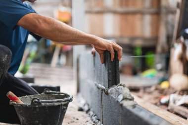 Adam duvara tuğla örüyor. Tuğlalar siyah ve adam mavi bir gömlek giyiyor.