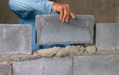 Adam duvara tuğla örüyor. Tuğlalar gri ve adam mavi bir gömlek giyiyor.