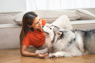 Kadın ve köpeği yerde birlikte yatıyorlar. Kadın turuncu bir gömlek giyiyor ve köpek bir Husky. Birbirlerine eşlik etmek hoşlarına gidiyor gibi. Kadın da köpeği okşuyor.