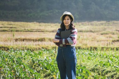 Şapka takan ve mavi tulumlu bir kadın yeşil bitki tarlasında duruyor. Elinde bir tablet tutuyor.