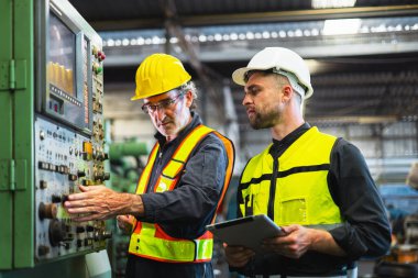Güvenlik teçhizatlı iki adam bir makine üzerinde çalışıyor. Onlardan biri bir tablet tutuyor. Sahne ciddi ve odaklı.
