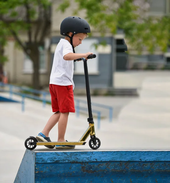 child rides a Kick scooter. playground for riding on Kick scooter. boy in a white T-shirt , hardhat and shorts riding a scooter in skatepark. young novice athlete spends free time in extreme sports.