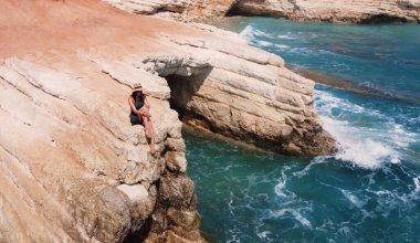 Beautiful girl sitting on a high rock and looking out to sea. top view. Girl in Hat on the edge of cliff. blue sea and high cliffs. Seascape. Girl at sunset. Sea tour. Blue sea, rocks. Cyprus