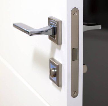 Modern ve minimalist tasarımı sergileyen beyaz bir kapının üzerindeki paslanmaz çelik kapı tokmağının detaylı görüntüsü. Manyetik kilidi aç. Gümüş saplı beyaz bir kapı ve kilidi var..