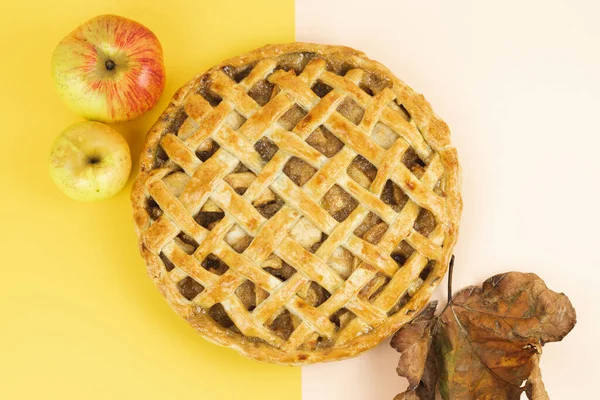 Baked apple pie with trellis decoration on a background of two warm colors. Apples and dried leaves. Copy space. Top view.