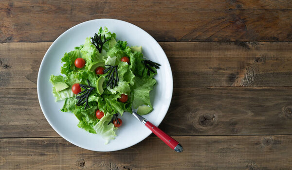 White plate of mixed salad with seaweed, tomatoes, lettuce and avocado. Wood background. Copy space. Top view.