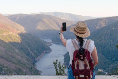 Sırt çantalı ve şapkalı bir kadın dağ manzarasında selfie çekiyor. Navia nehri, Salime rezervuarı. Paicega. Boşluğu kopyala.