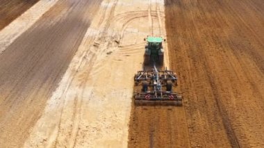 Tractor with harrow system plowing ground on cultivated farm field, pillar of dust trails behind, preparing soil for planting new crop, agriculture concept, top view.