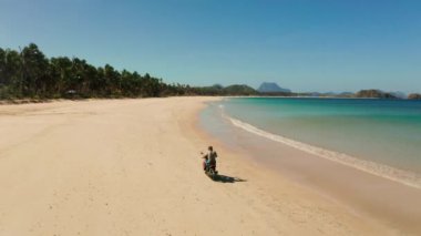 Plajda motosiklet süren şapkalı adamın hava görüntüsü. Adam güneşli bir günde scooter kullanıyor. Okyanus sahilinde motorsikletle dalgalara karşı. Nacpan, El Nido, Palawan, Filipinler.