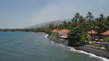 Havuzu, güneşli yatakları, deniz kenarında palmiye ağaçları olan lüks bir otel. Siyah kumlu sahil şeridi, palmiye ağaçları, otel, tropikal tatil köyü. Bali, Endonezya, seyahat konsepti. hava görüntüleri
