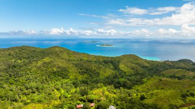 Mavi gökyüzünün altında durgun bir turkuaz denize doğru uzanan yuvarlak yeşil tepelerin manzarası. Praslin, Seyşeller.