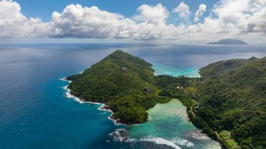 Kayalık kıyıları ve sık orman bitkilerinin yer aldığı yeşil adalı geniş bir gök mavisi okyanus. Seyşeller, Mahe.