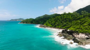 Dalgalar, yoğun, yeşil tropikal bitki örtüsüyle çerçevelenmiş engebeli kayalara çarpar. Seyşeller, Mahe.