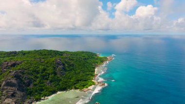 Turkuaz sular yoğun yeşil tepelerin altındaki kayalık kıyılara çarpar. La Digue, Seyşeller.