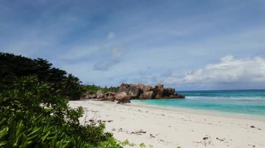 Turkuaz okyanus dalgaları, açık ve parlak bir gökyüzünün altında, beyaz kumsalla nazikçe buluşuyor. Seyşeller, La Digue. Anse Komandoları.