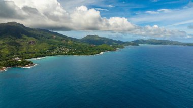 Yeşil kıyı şeridi, parçalı bulutlu bir gökyüzünün altında turkuaz Hint Okyanusu ile buluşur. Seyşeller, Mahe.