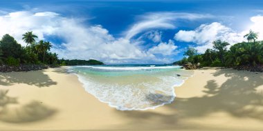 Yumuşak dalgalar, palmiye ağaçlarıyla gölgelenmiş ve güneşli bir gökyüzünün altında büyük kayalarla çevrili altın kumlu bir plaja yuvarlanıyor. Anse Takamaka Sahili, Seyşeller. VR 360.