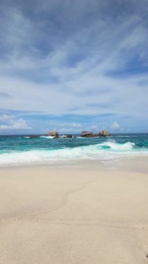 Dalgalar, berrak bir gökyüzünün altında, canlı turkuaz sularla beyaz kumlu bir sahilde kayalara çarpar. Seyşeller, Mahe. Anse Boileau.