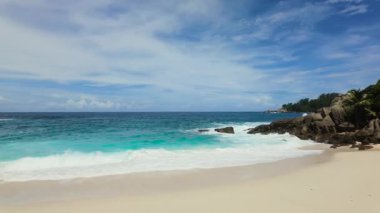 Temiz bir gökyüzünün altında dağınık kayalarla el değmemiş beyaz kumlu bir plajda hafif turkuaz dalgalar. Seyşeller, Mahe. Anse Boileau.