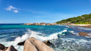 Güçlü okyanus dalgaları gür orman bitki örtüsü ve açık mavi gökyüzü ile kayalık kıyıya çarpar. Seyşeller, La Digue.