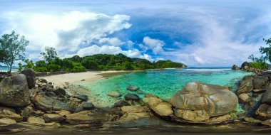 Kristal berrak sular, parlak bir gökyüzünün altında yeşil bir zemin ile küçük bir plajdaki kayaları çevrelemektedir. Port Glaud Gölü. Seyşeller. VR 360.