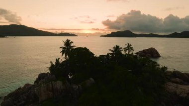 Güneş uzak adaların ardında batarken sakin sulara bakan palmiye ağaçları ve kayaların olduğu tropik bir manzara. Praslin, Seyşeller.