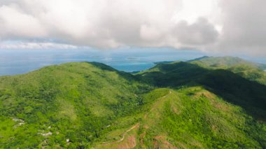 Yemyeşil tepeler, ufukta küçük adalar olan kayan bulutlardan oluşan bir gökyüzünün altında okyanusa doğru çağlar. Praslin, Seyşeller.