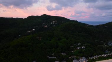 Dağılmış evleri ve pembe bir gökyüzünün altında kıyı manzarası olan bereketli bir dağ manzarası. Seyşeller, Mahe.