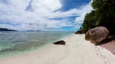 Büyük granit kayaları, turkuaz okyanusu ve parlak mavi gökyüzünün altında yemyeşil palmiye ağaçları olan beyaz kumsal. Seyşeller, La Digue.