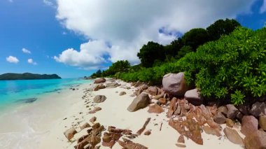 Yeşil bitki örtüsü, granit kayalar ve parçalı bulutlu bir gökyüzünün altında turkuaz bir deniz bulunan kıyı manzarası. Anse Takamaka. Seyşeller, Praslin.