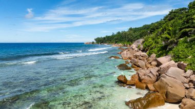 Tropikal bitki örtüsü temiz mavi su ve dağınık kayalarla kıyı şeridi oluşturur. La Digue, Seyşeller.