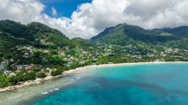 Kumlu bir sahil ve turkuaz su manzaralı dağınık evleri olan yeşil tepeler. Seyşeller, Mahe. Beau Vallon Plajı.