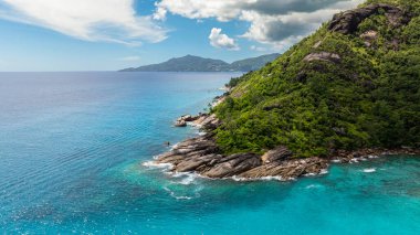 Turkuaz okyanus suyu kayalık bir yamaç boyunca yeşil bitki örtüsüyle kaplı, uzak dağlarla kaplı. Seyşeller, Mahe.