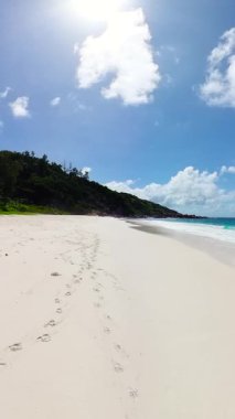 Yumuşak beyaz kumdaki ayak izleri, yemyeşil bitki örtüsü ve açık mavi gökyüzü ile tropik bir sahil boyunca uzanıyor. Küçük Anse. Seyşeller, La Digue.