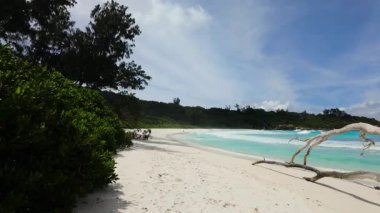 Dalgalar, yemyeşil bitki örtüsüyle çevrili beyaz kumsalda yavaşça kıyıya vurur. Seyşeller, La Digue. Anse Komandoları.