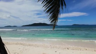 Nazik turkuaz dalgalar kumlu sahilde, parlak bir gökyüzünün altında uzak bir adaya vurur. Seyşeller, La Digue.