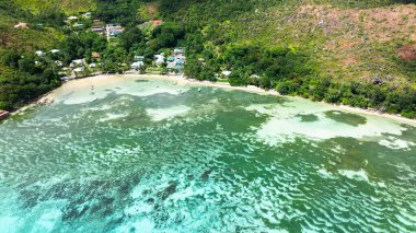 Arka planda yemyeşil tepelerin yükseldiği kıyı köyü yakınlarındaki sığ ve berrak suların havadan görünüşü. Praslin, Seyşeller.