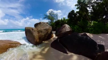 Okyanus dalgalarının parlak bir gökyüzünün altında dalgalandığı kumlu bir sahil boyunca devasa kayalar. Seyşeller, Mahe. Anse Takamaka.