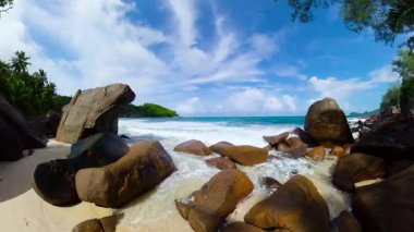 Okyanus dalgalarının parlak mavi bir gökyüzünün altına düştüğü kumlu bir sahilde büyük kayalar. Seyşeller, Mahe. Anse Takamaka.