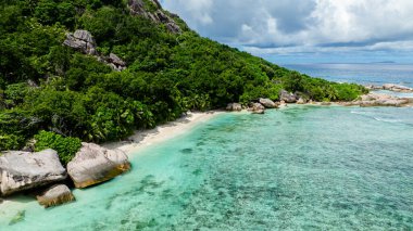 Yeşillik ve büyük kaya oluşumlarıyla çevrili, kumlu bir plajda kristal berrak turkuaz su. Seyşeller, La Digue.