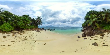 Dağınık kayalar ve turkuaz sularla çevrili, bulutlu bir gökyüzünün altında yemyeşil tropikal ağaçlarla çevrili sessiz bir sahil. Anse Bougainville Sahili, Seyşeller. 360 görünüm.